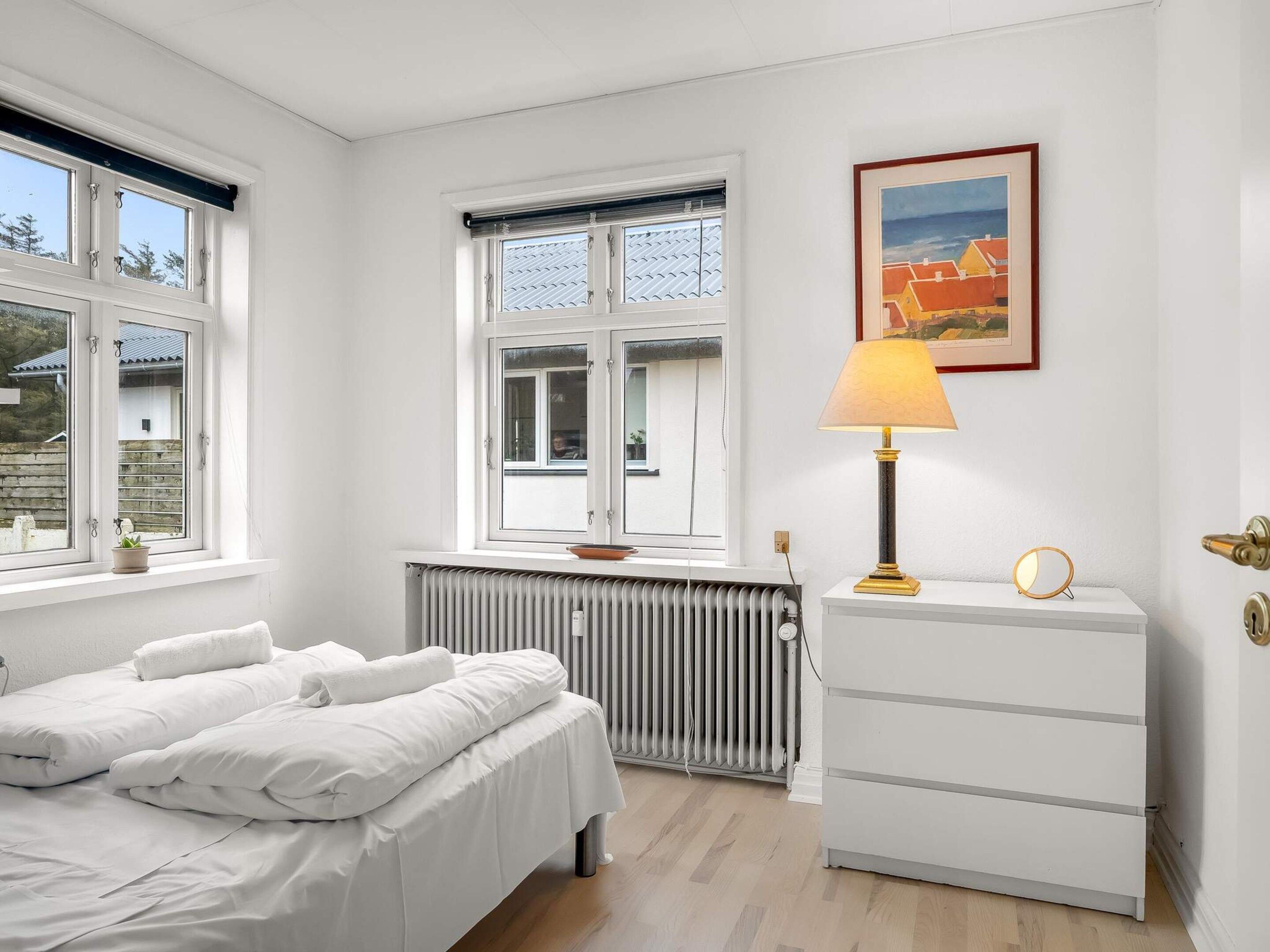 A bedroom with a bed, a radiator, a dresser, a lamp, and a window with a view.