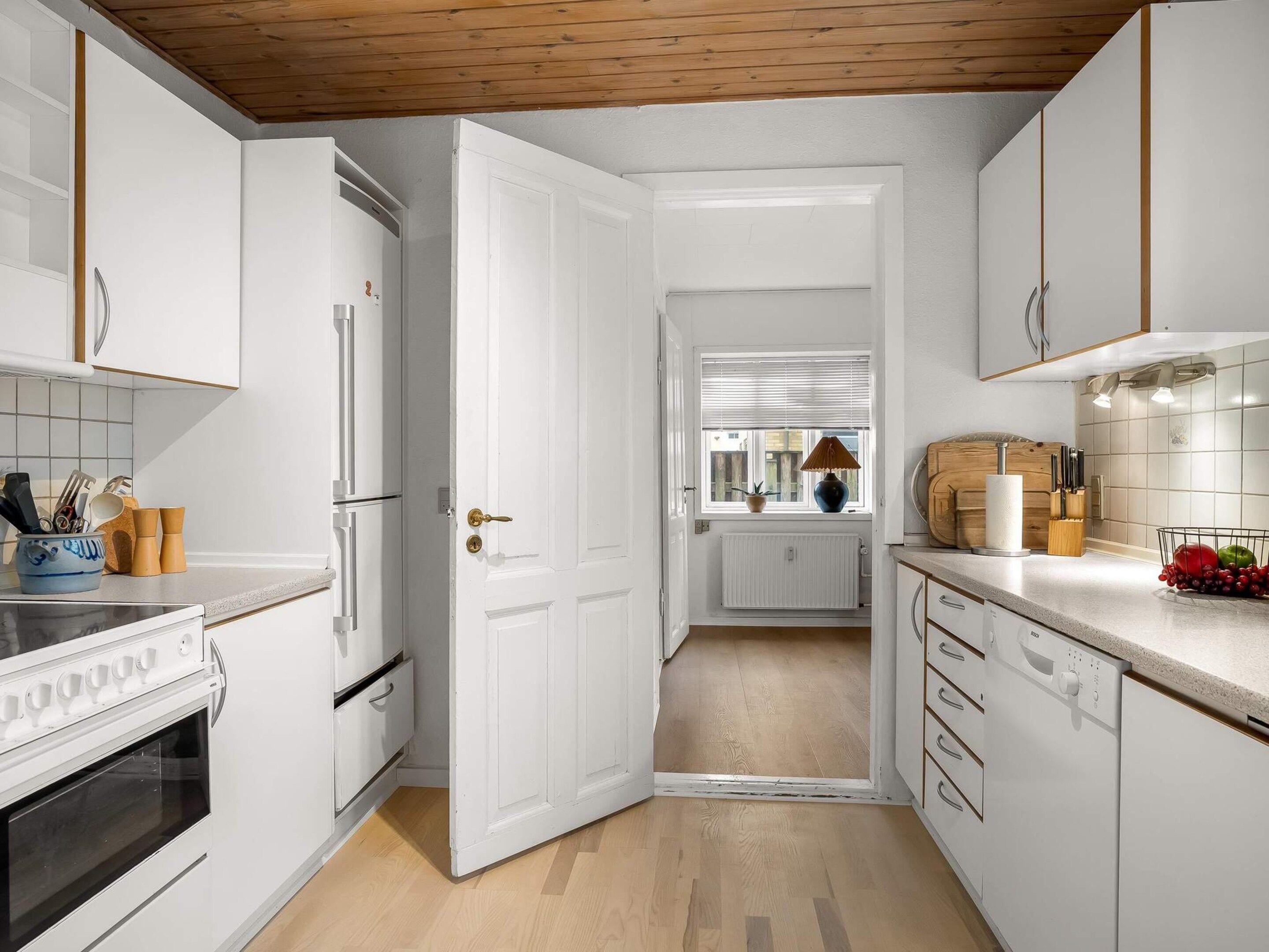 A modern kitchen with white cabinetry, a built-in oven, a refrigerator, and a window with blinds.