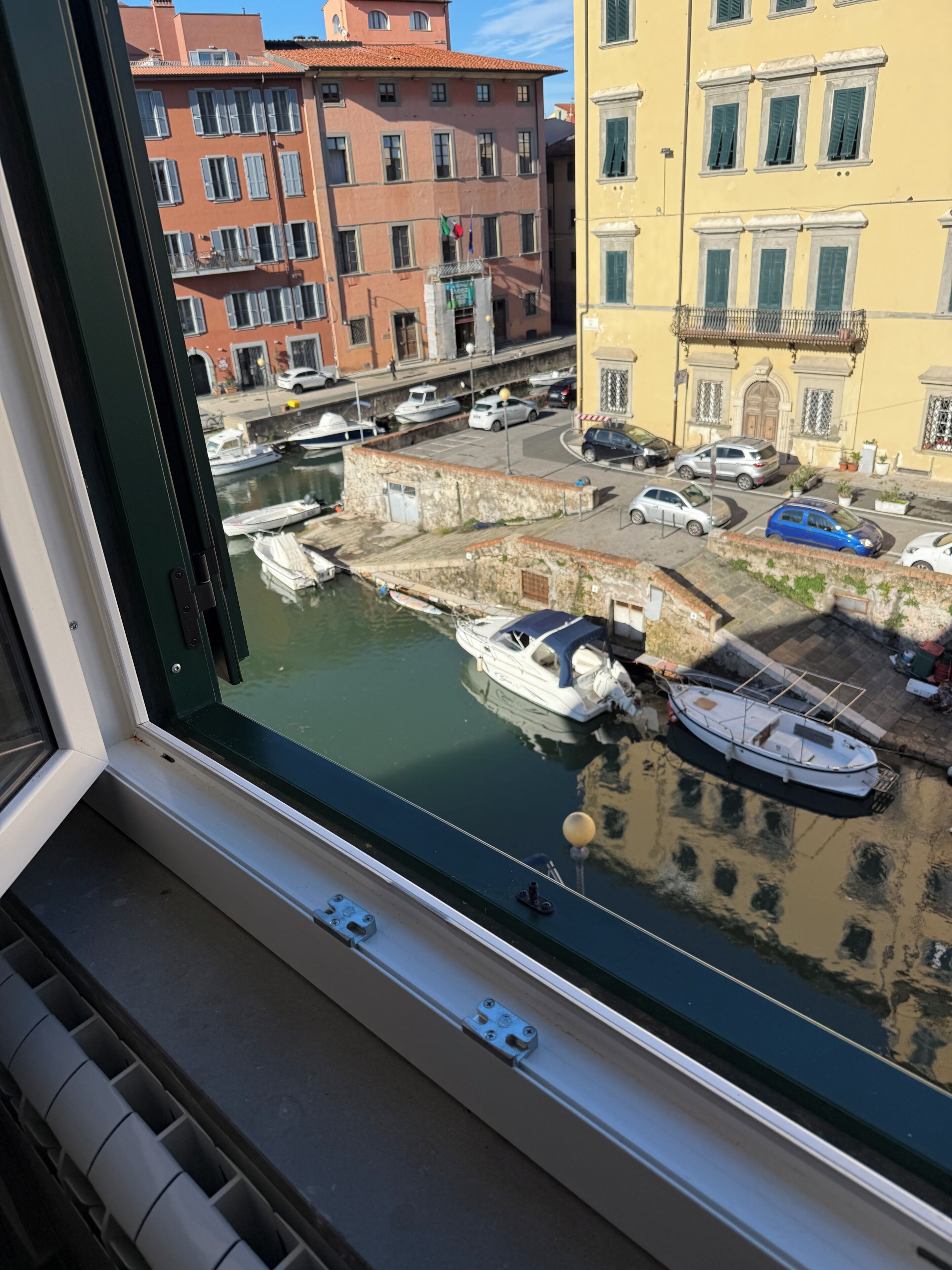 Vista a través de una ventana de un puerto deportivo con barcos amarrados, edificios al fondo y un cielo despejado.