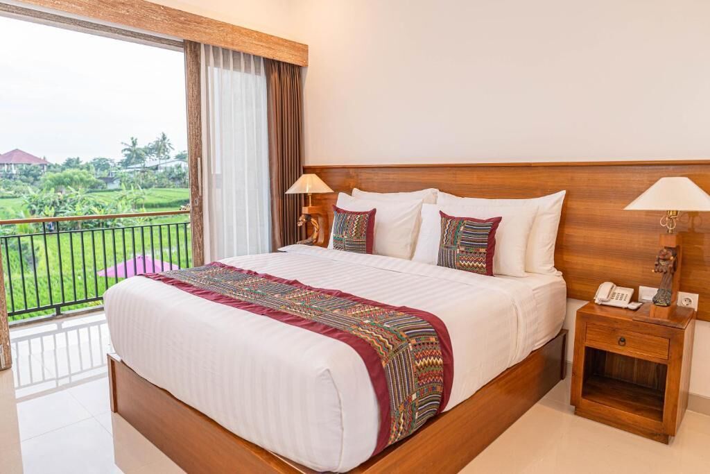 Habitación de hotel con una cama grande, dos mesitas de noche, un balcón con vistas a zonas verdes y un cabecero de madera.