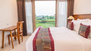 A hotel room with a bed, a desk, a chair, and a view of a green landscape through large windows.