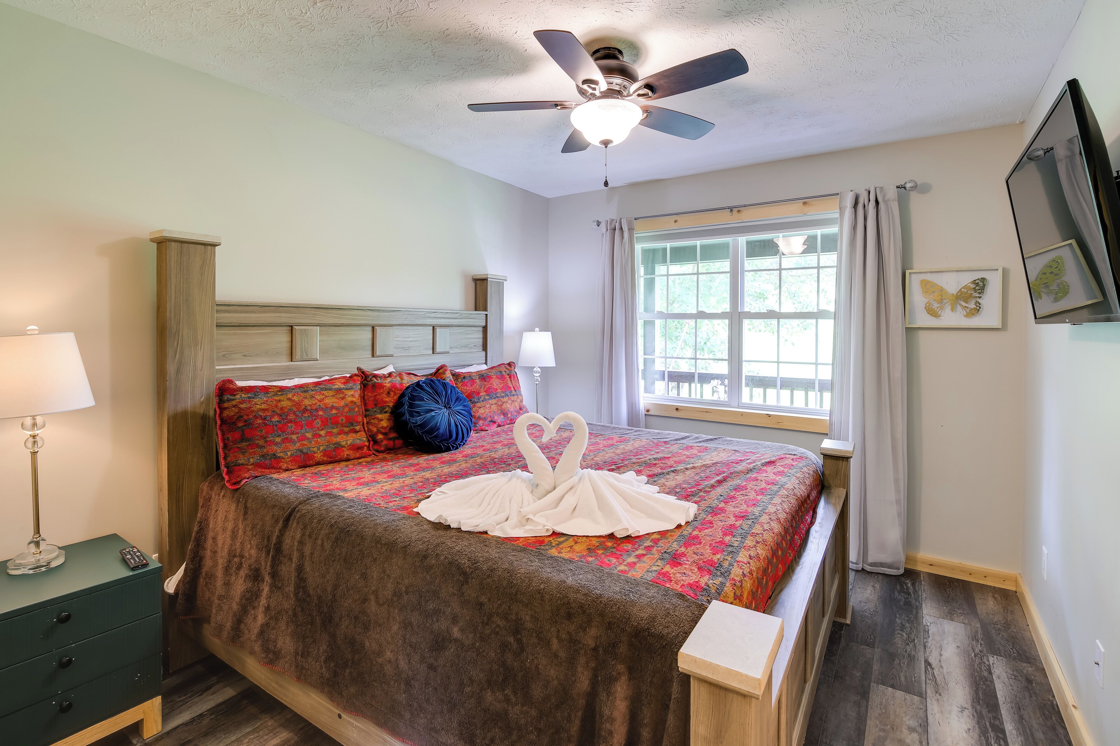 Un lit bien fait, agrémenté d’un coussin décoratif en forme de cygne, un ventilateur de plafond, une lampe de chevet et une fenêtre avec des rideaux.