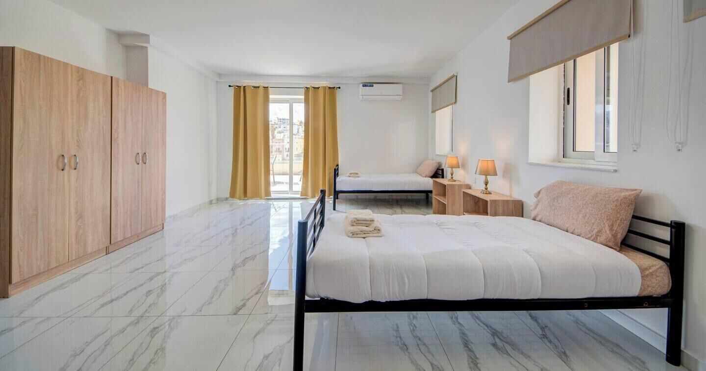 A bedroom with a bed, a wardrobe, a nightstand with lamps, and a window with curtains.