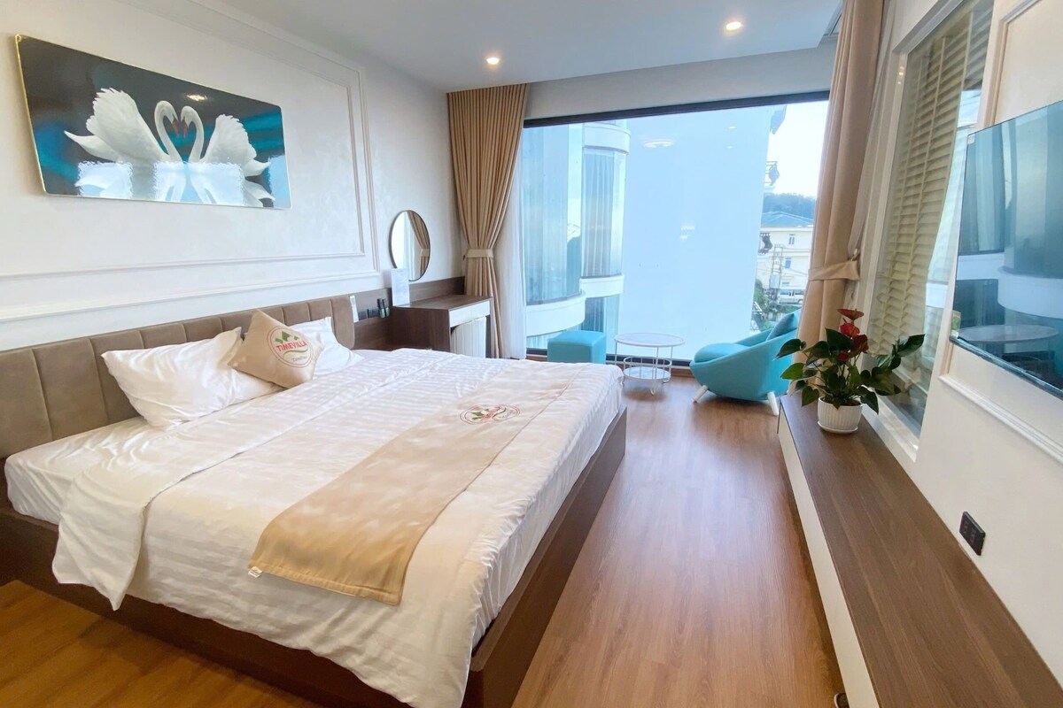 Een hotelkamer met een bed, nachtkastje, spiegel, raam met gordijnen en een balkon met stoel.