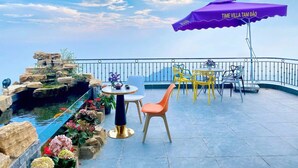 Una terraza con vistas, con una mesa y sillas, un paraguas morado, y un pequeño jardín con rocas y flores.