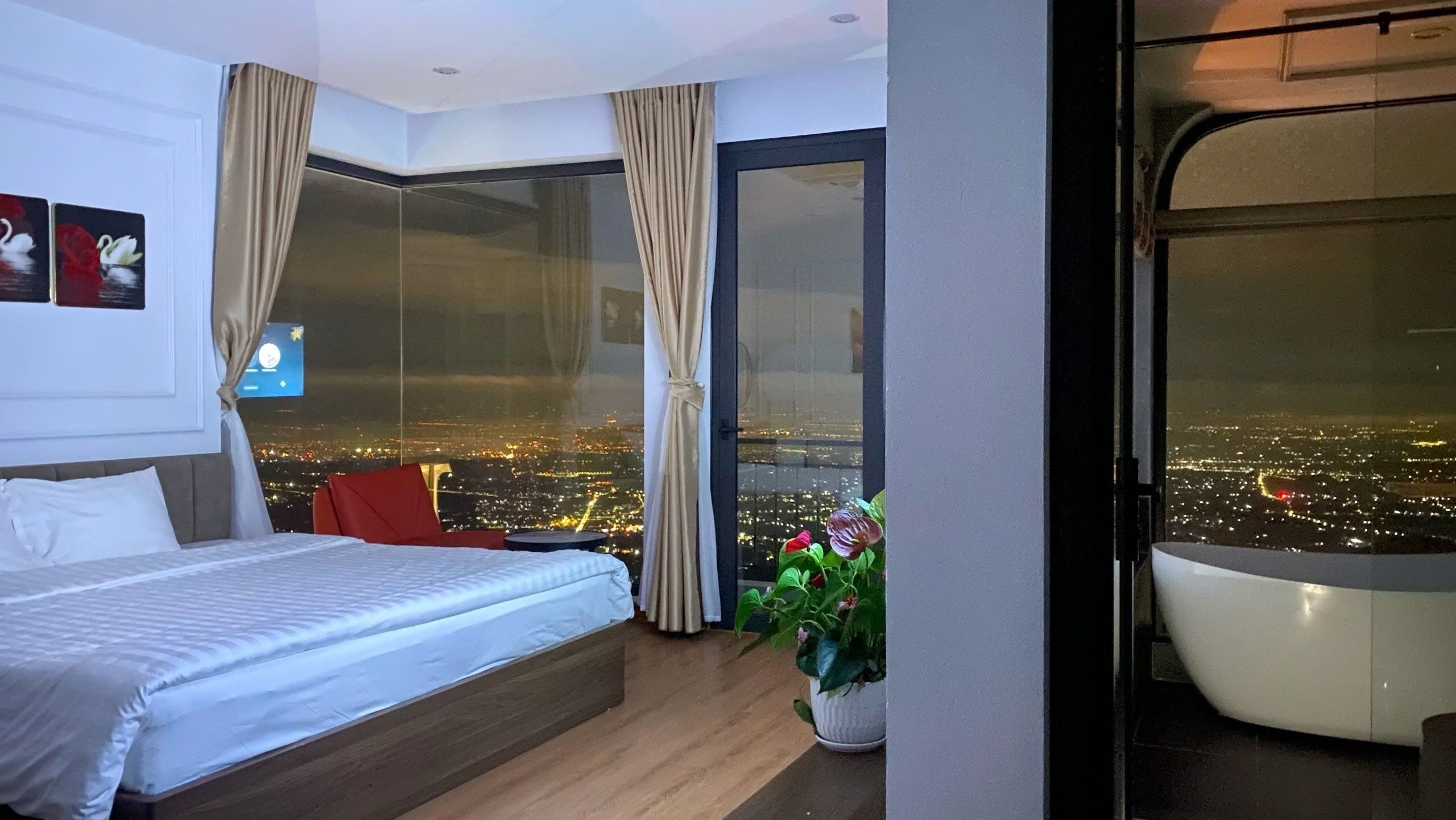 A modern hotel room with a bed, a red chair, a potted plant, and a large window offering a city view.