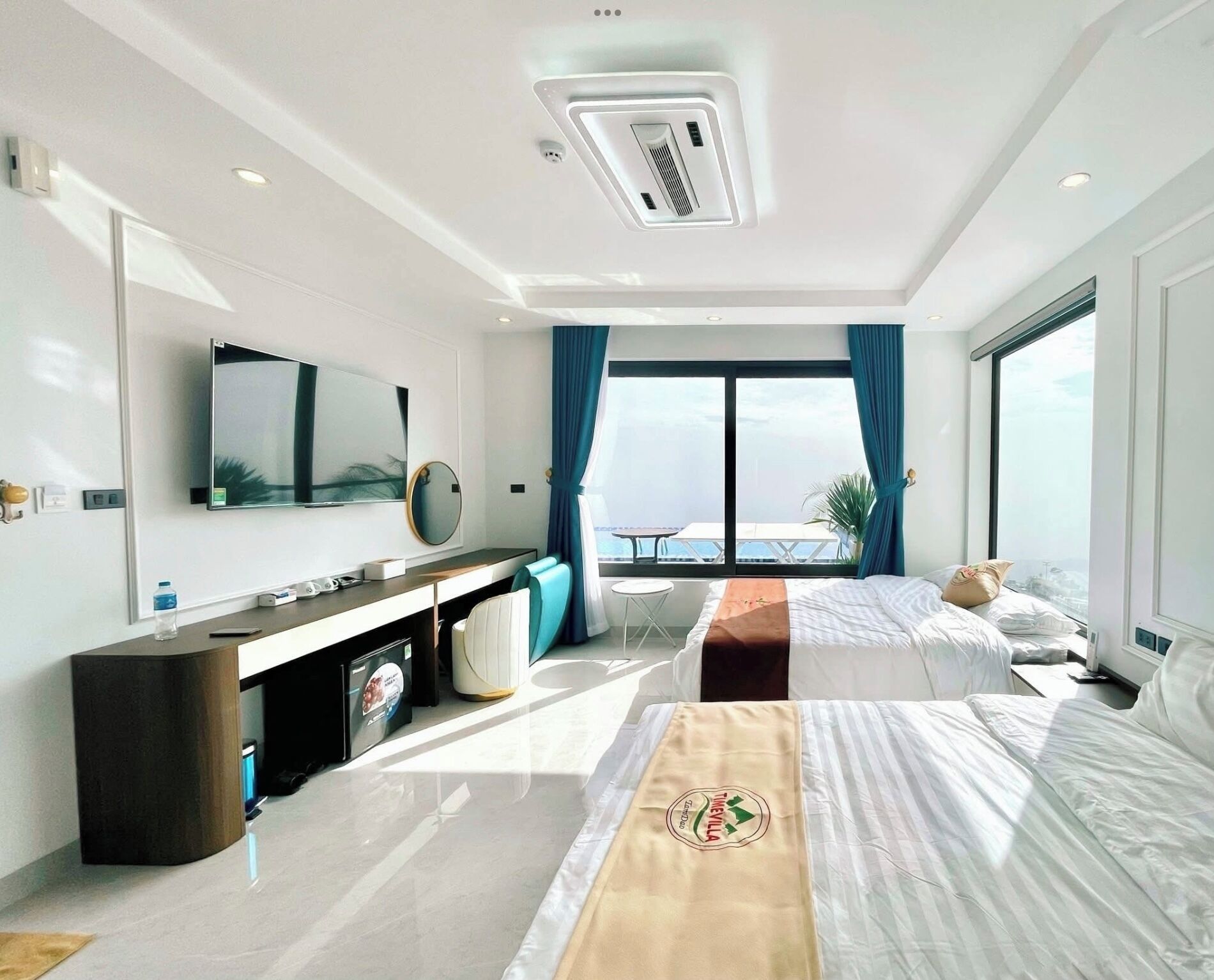 A modern hotel room with a large bed, a desk with a computer, a TV, and a view of the sea.