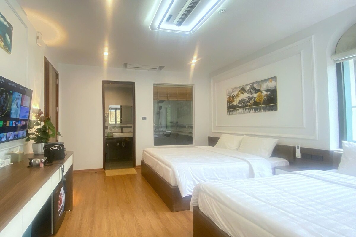 A hotel room with a wooden floor, two beds, a TV, a painting, and a bathroom visible through an open door.