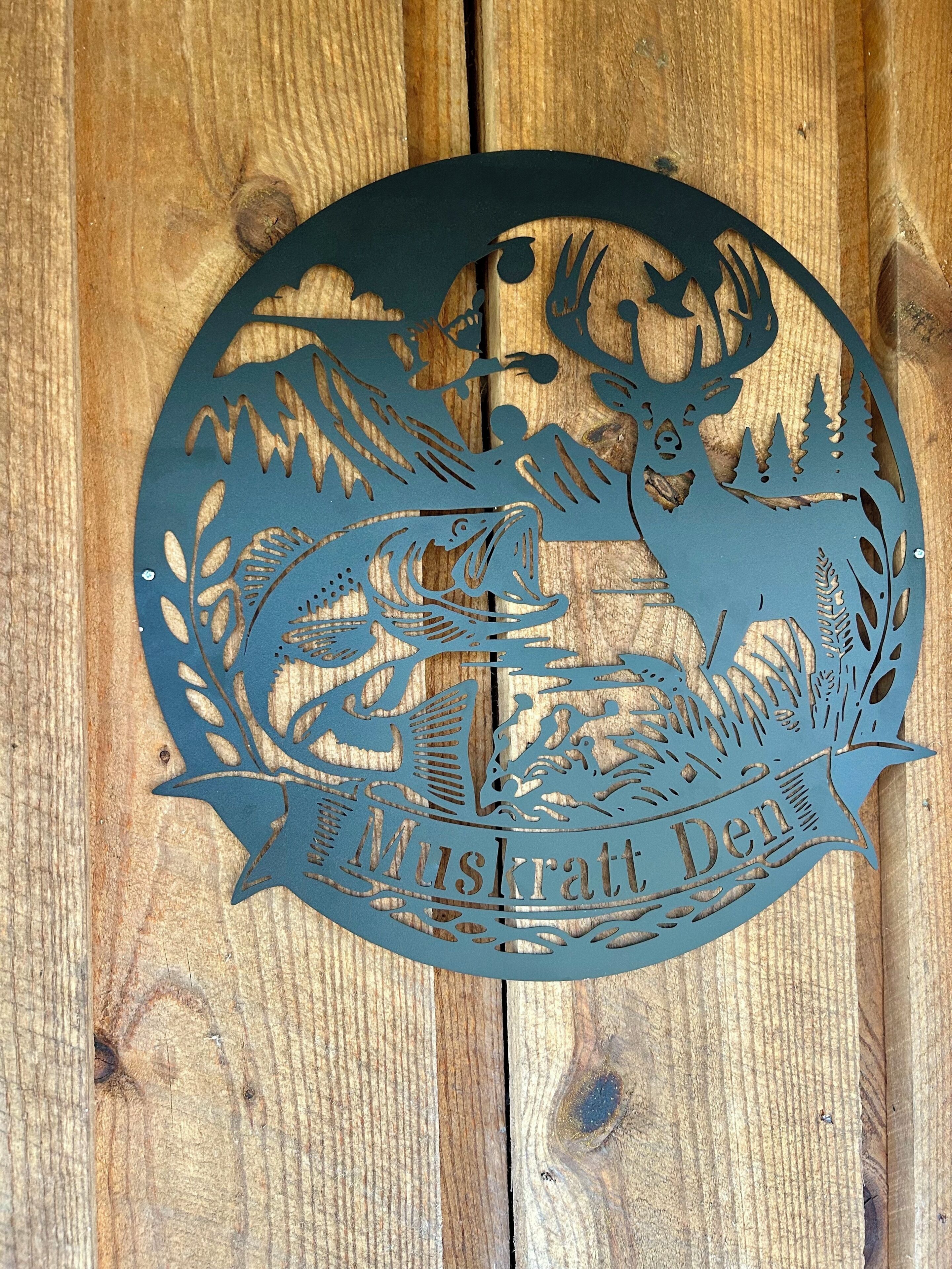 Pared de madera con un recorte metálico que muestra un ciervo y follaje, con el texto 'Muskratten Den'.
