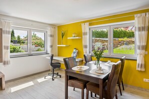 Et spiseområde med et bord dækket til fire personer, stole, en vase med blomster og et vindue med gardiner.