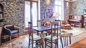 Une pièce aux murs de pierre, comprenant une table à manger, des chaises, un piano et une bibliothèque.