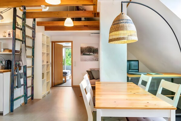 Eine moderne Küche mit Holzbalken, einem Essbereich mit Holztisch und weißen Stühlen sowie einer großen, gewebten Pendelleuchte.