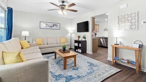 Ein modernes Wohnzimmer mit einer Couch, einem Couchtisch und einem Fernseher. Ein Deckenventilator ist vorhanden, im Hintergrund befindet sich eine Küche und ein Teppich mit Muster.