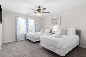 Habitación de hotel con dos camas, ventilador de techo, ventana con cortinas estampadas y una obra de arte colgada en la pared.