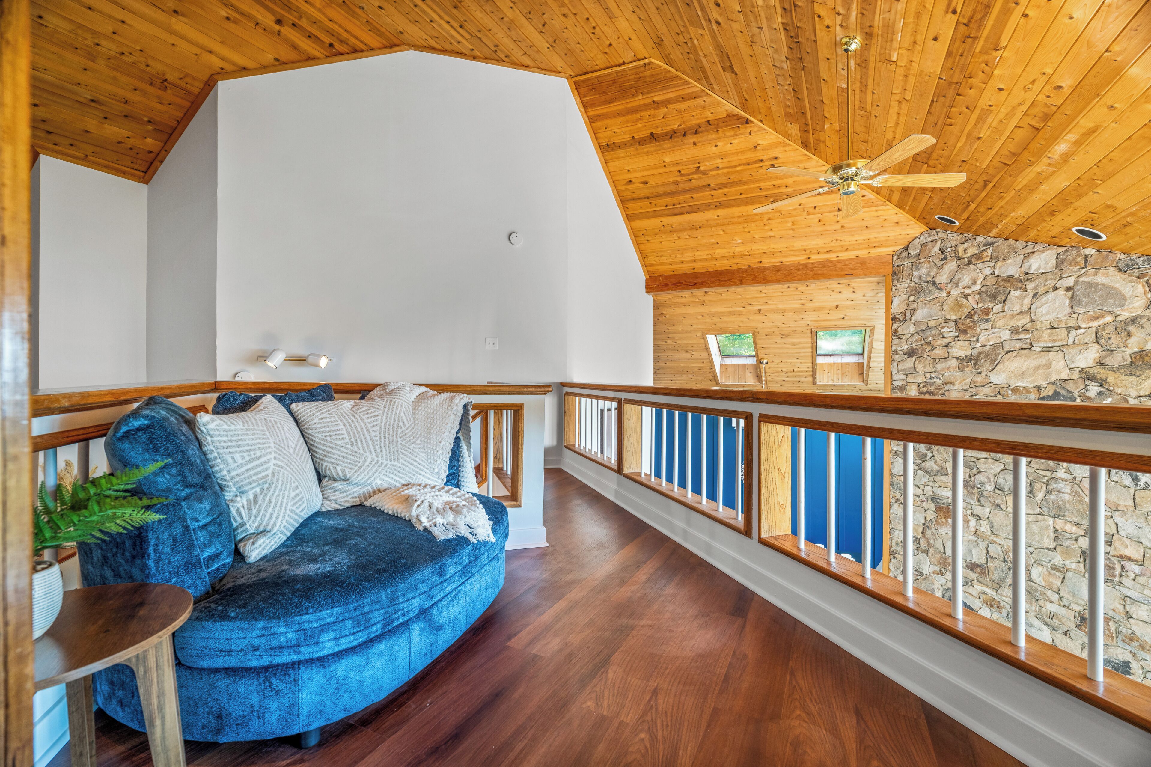 A spacious room with a wooden ceiling, a stone accent wall, a blue sofa, and a balcony with a railing.