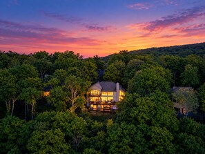 A two-story house surrounded by dense forest, with a visible deck and multiple windows. - Wildbriar Lodge • Luxe 6BR, 6,100 SqFt • Theater, 2xGameRoom, Karaoke, Sleeps 22 (Nellysford)