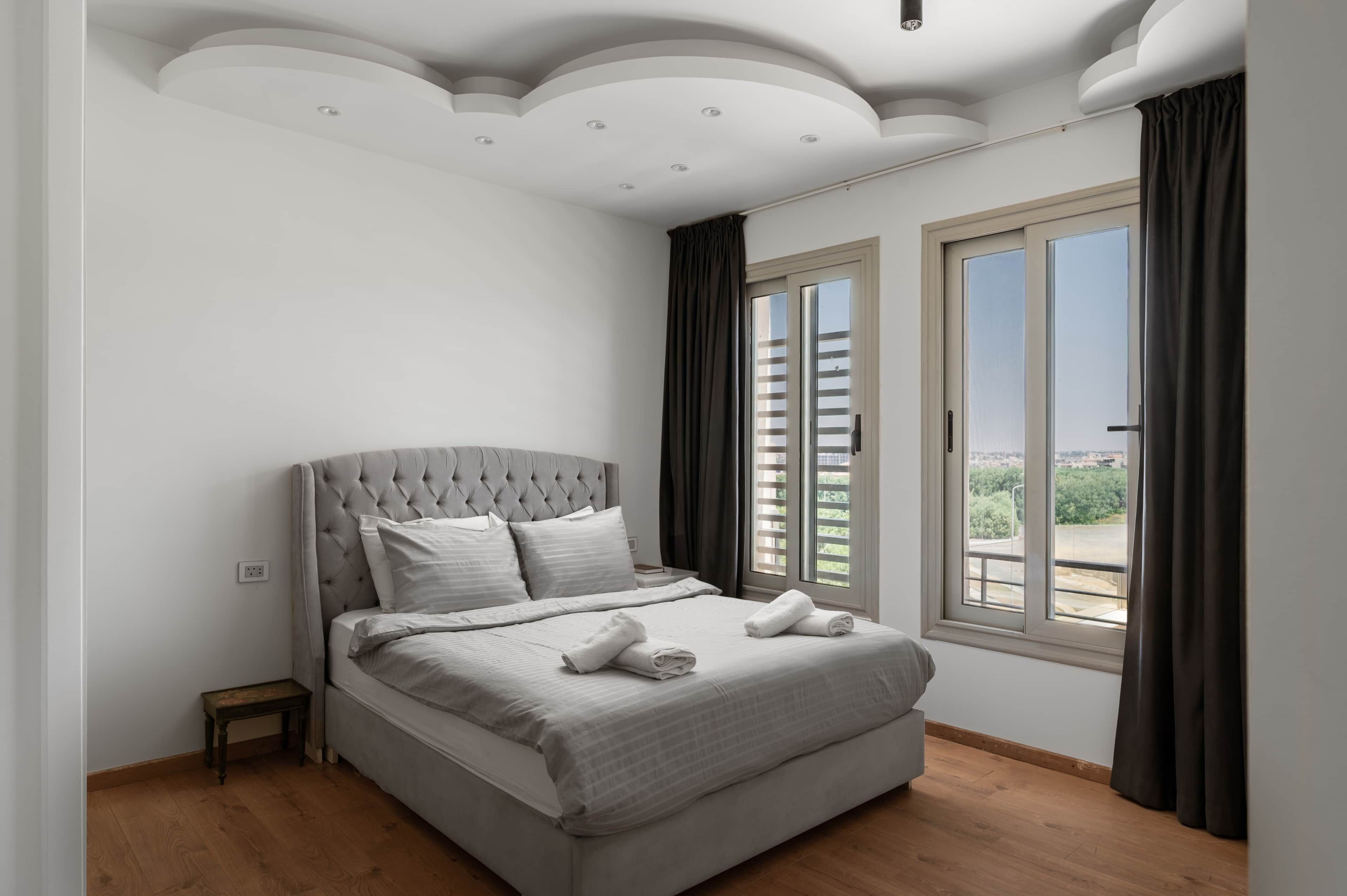 A bedroom with a tufted headboard, a bed with white linens, a small bedside table, a window with curtains, and a view of greenery outside.