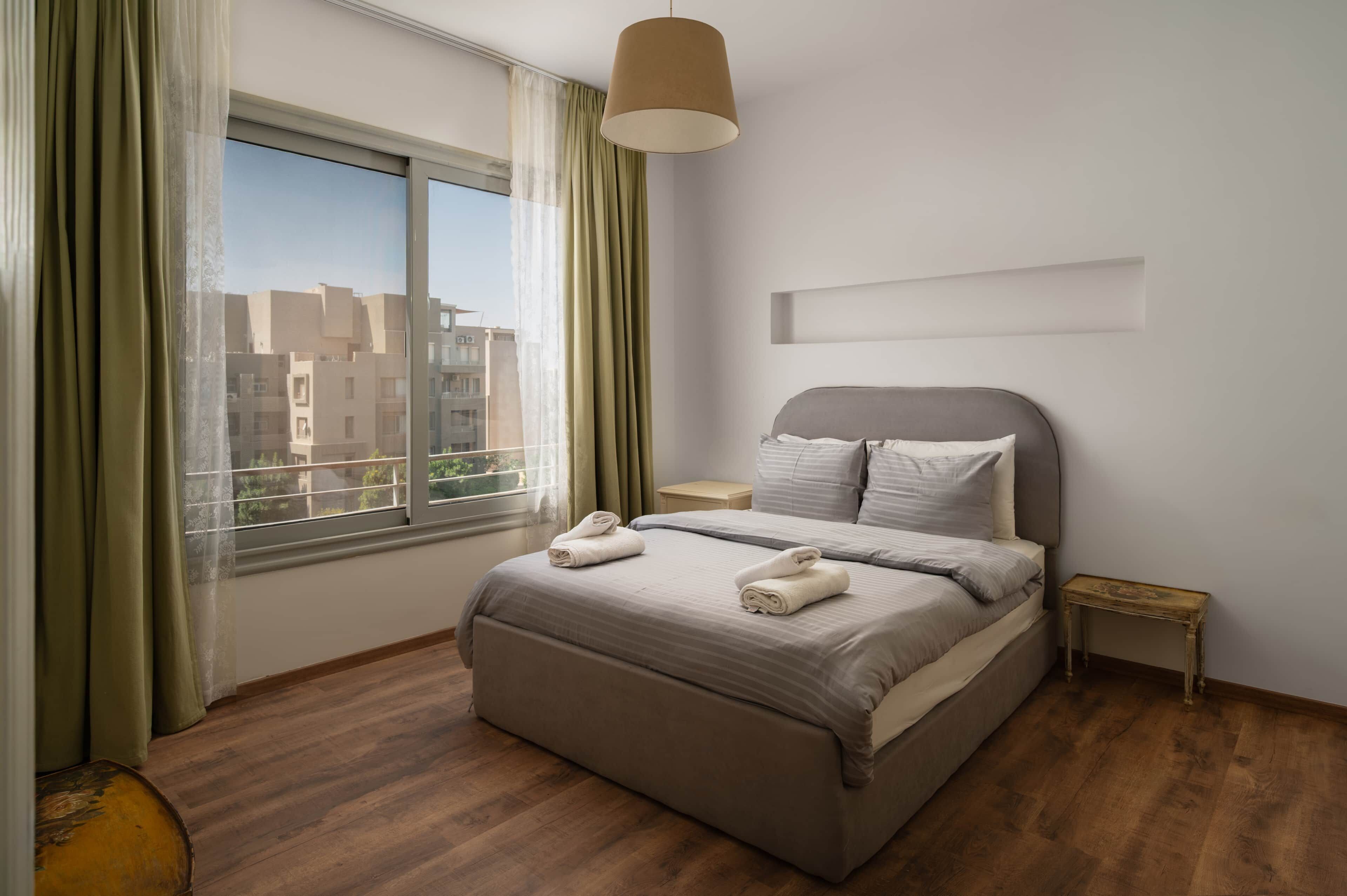 Une chambre d’hôtel avec une grande fenêtre, un lit avec des oreillers, une table de chevet et une lampe.