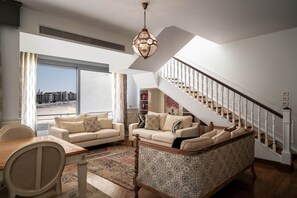 A spacious living room with a high ceiling, a staircase, and a large window overlooking a cityscape. - The White Pent - Palm Hills I Nesty Hospitality (New Cairo City)