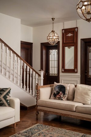 Eine Treppe mit weißen Balustraden führt zu einer dunklen Holztür, eine hängende Chandelieber und ein beiges Sofa mit einem floralen Kissen.