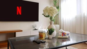 Ett vardagsrum med ett soffbord i glas, en tv-skärm som visar Netflix-logotypen, en vas med vita blommor, en liten krukväxt och en fjärrkontroll.