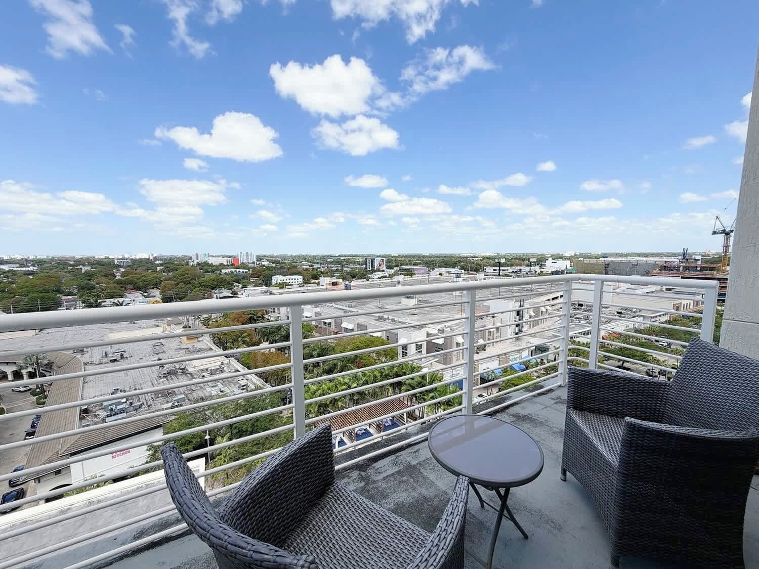 A balcony with wicker furniture, a small table, and a view of a cityscape with buildings and greenery.