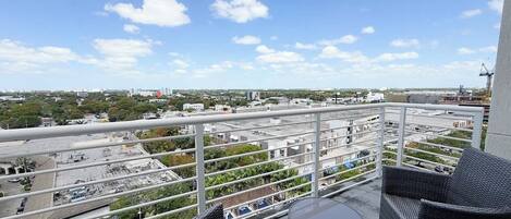 A balcony with wicker furniture, a small table, and a view of a cityscape with buildings and greenery.