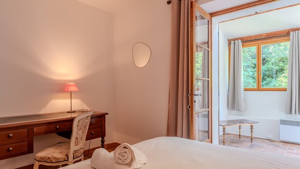 Une chambre à coucher comprenant un lit, un bureau, une chaise, une lampe et donnant sur l’extérieur.