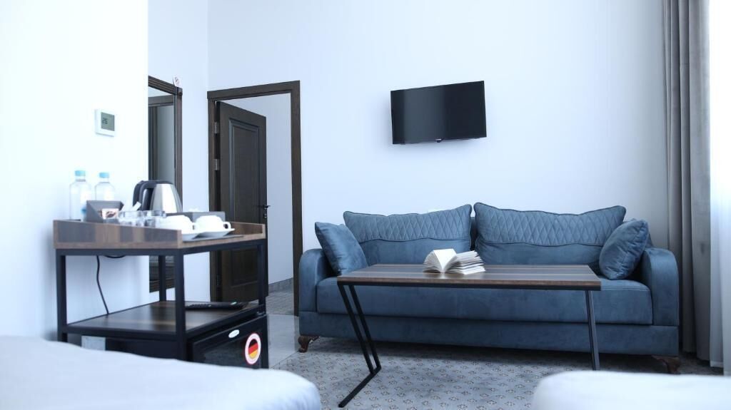 Ett modernt hotellrum med en blå soffa, ett litet bord med en bok, en TV som är monterad på väggen och ett skrivbord med en vattenkokare och vattenflaskor.