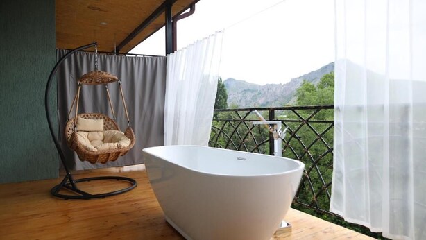 A room with a white bathtub, a wicker hanging chair, and a view of mountains through a balcony. - BMG Hotel  (Goris)