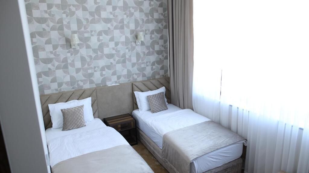Habitación de hotel con dos camas, cabecero, mesita de noche y ventana con cortinas.