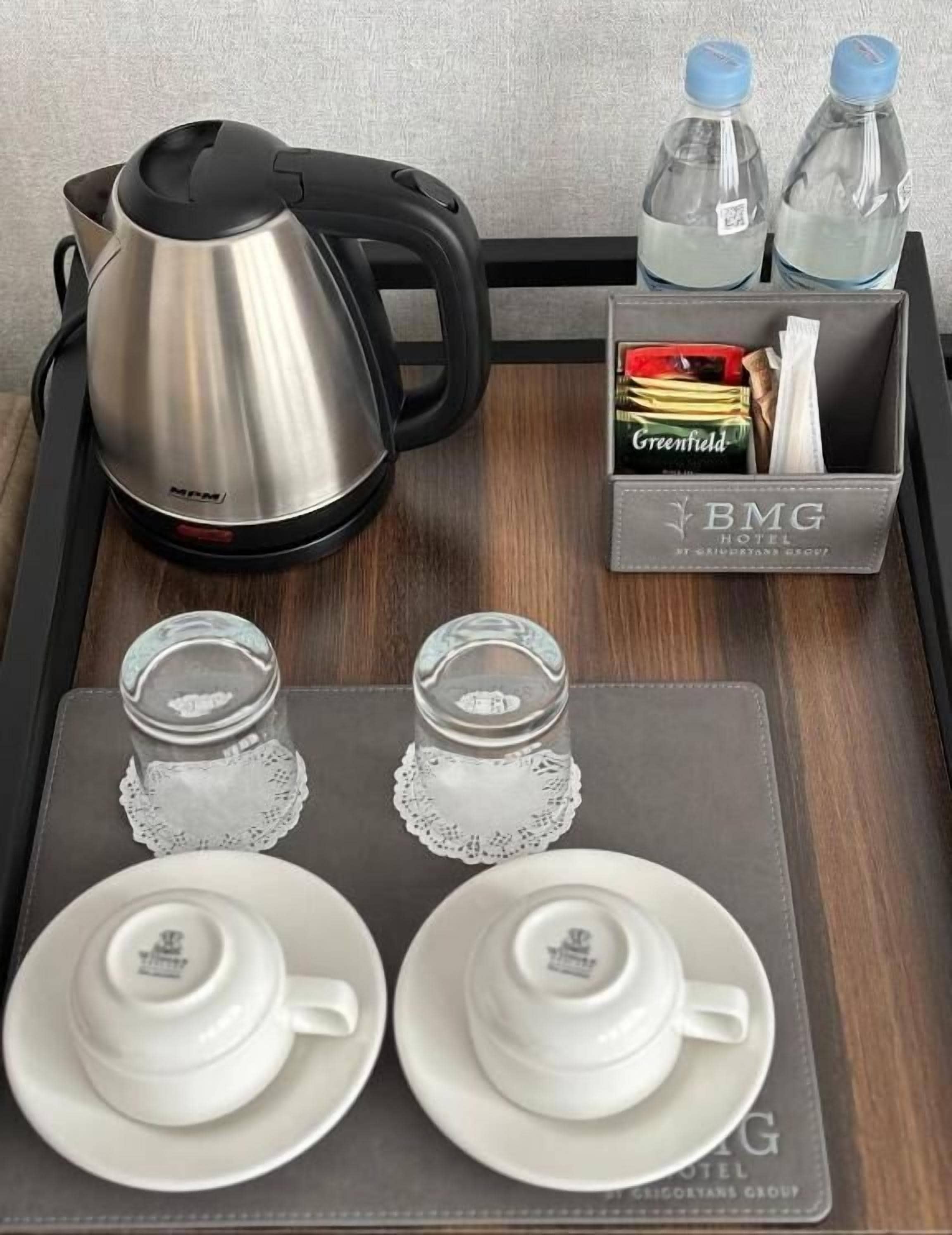 A hotel room tray with a kettle, water bottles, a box of Greenfield cigarettes, salt and pepper shakers, and two sets of white tea cups and saucers.