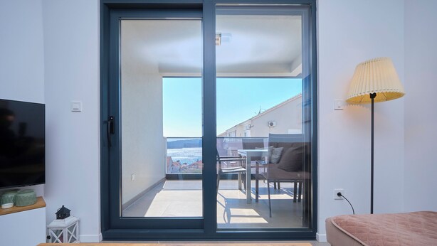 A modern living room with a sofa, a TV, a lamp, and a sliding glass door leading to an outdoor balcony with a table and chairs. - Ray Holiday Apartment (Okrug Gornji)