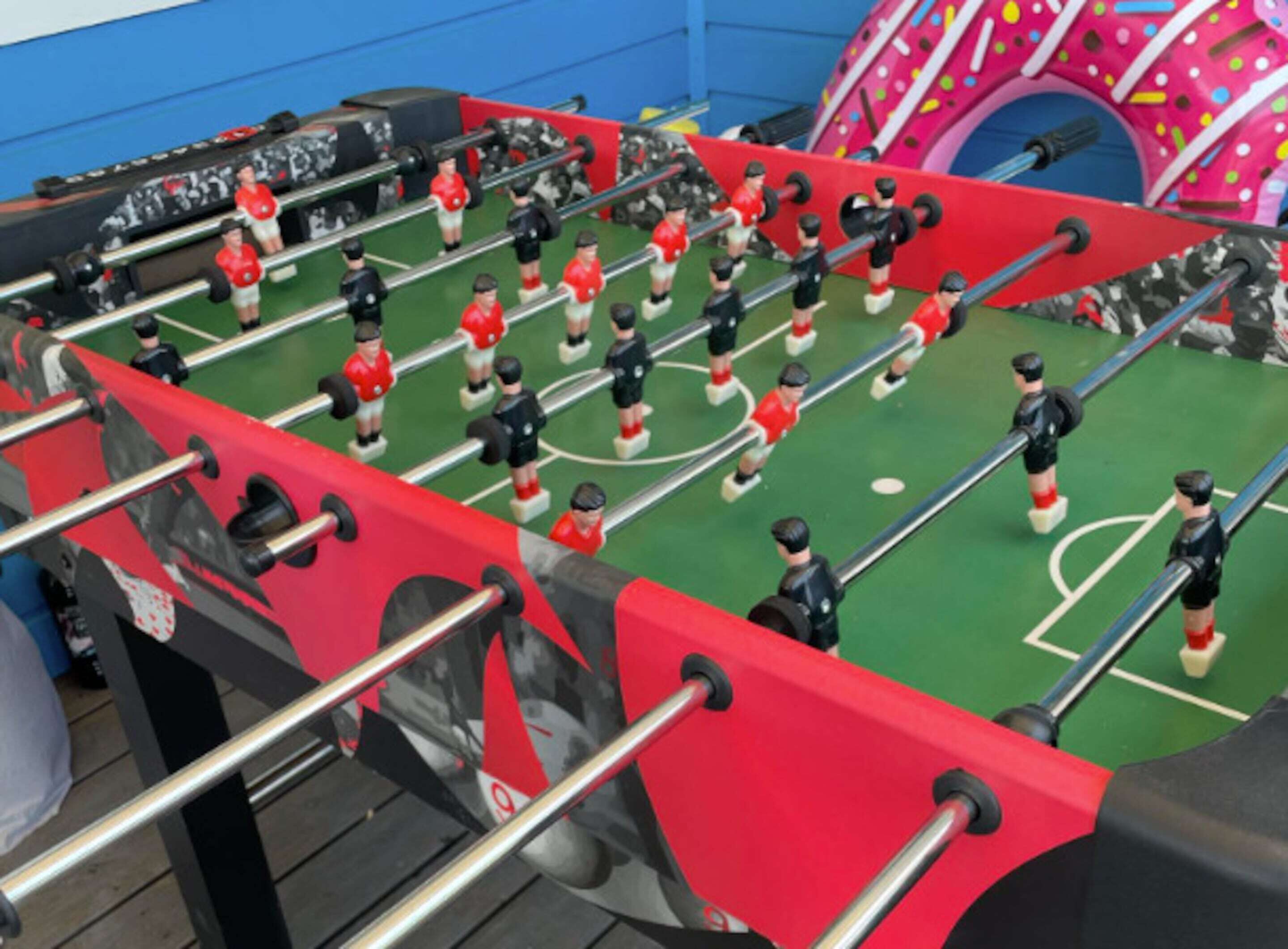 Une table de baby-foot avec des figurines rouges et noires, un tapis de jeu vert et un mur bleu en arrière-plan.