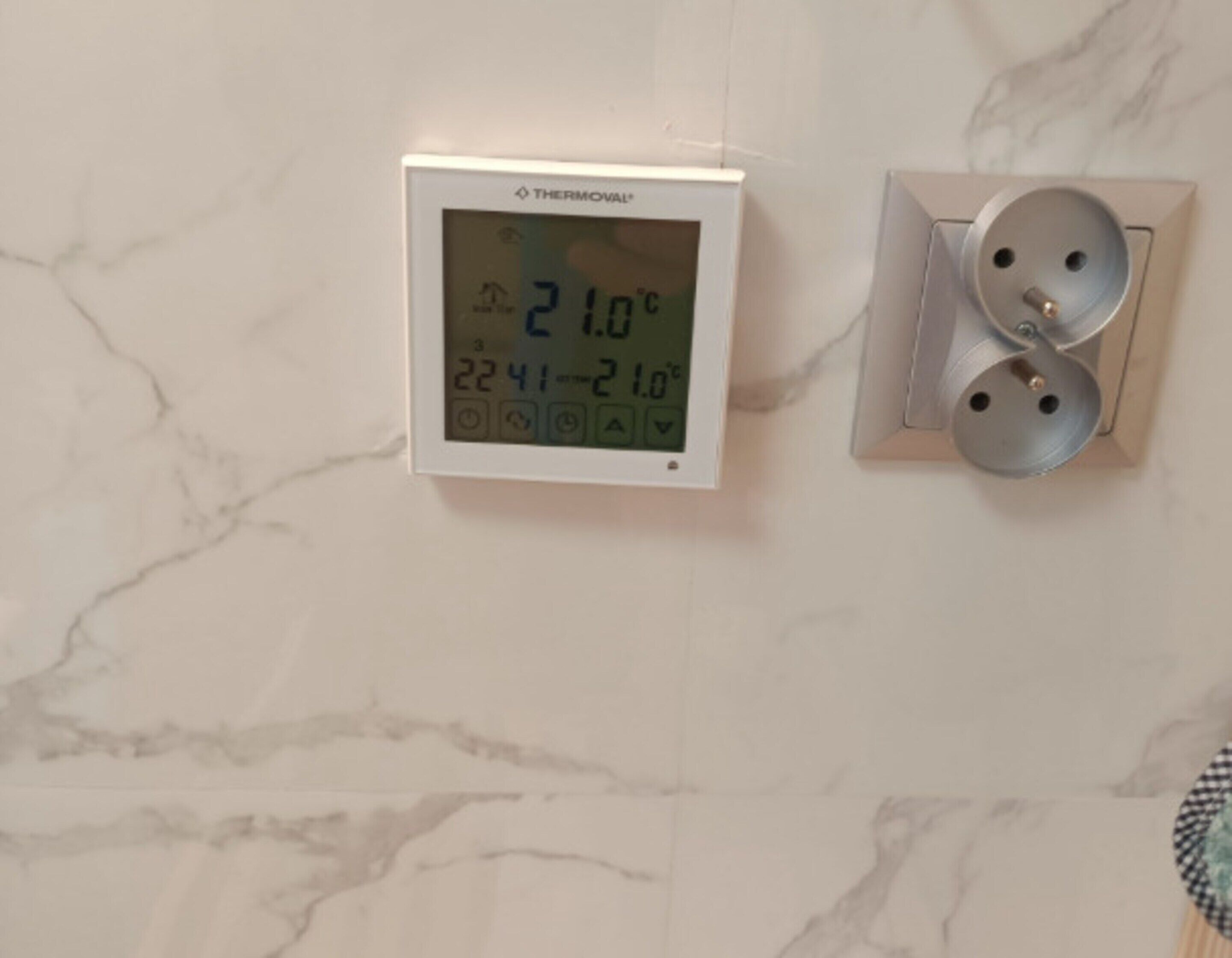 Termostato fijado a la pared que muestra una temperatura de 21.0°C, con un panel de control que tiene dos mandos circulares y un enchufe eléctrico visible.