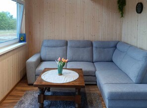 Un salón con un sofá gris, una mesa de centro de madera con un posavasos blanco, una alfombra azul y una ventana con vistas a la vegetación.