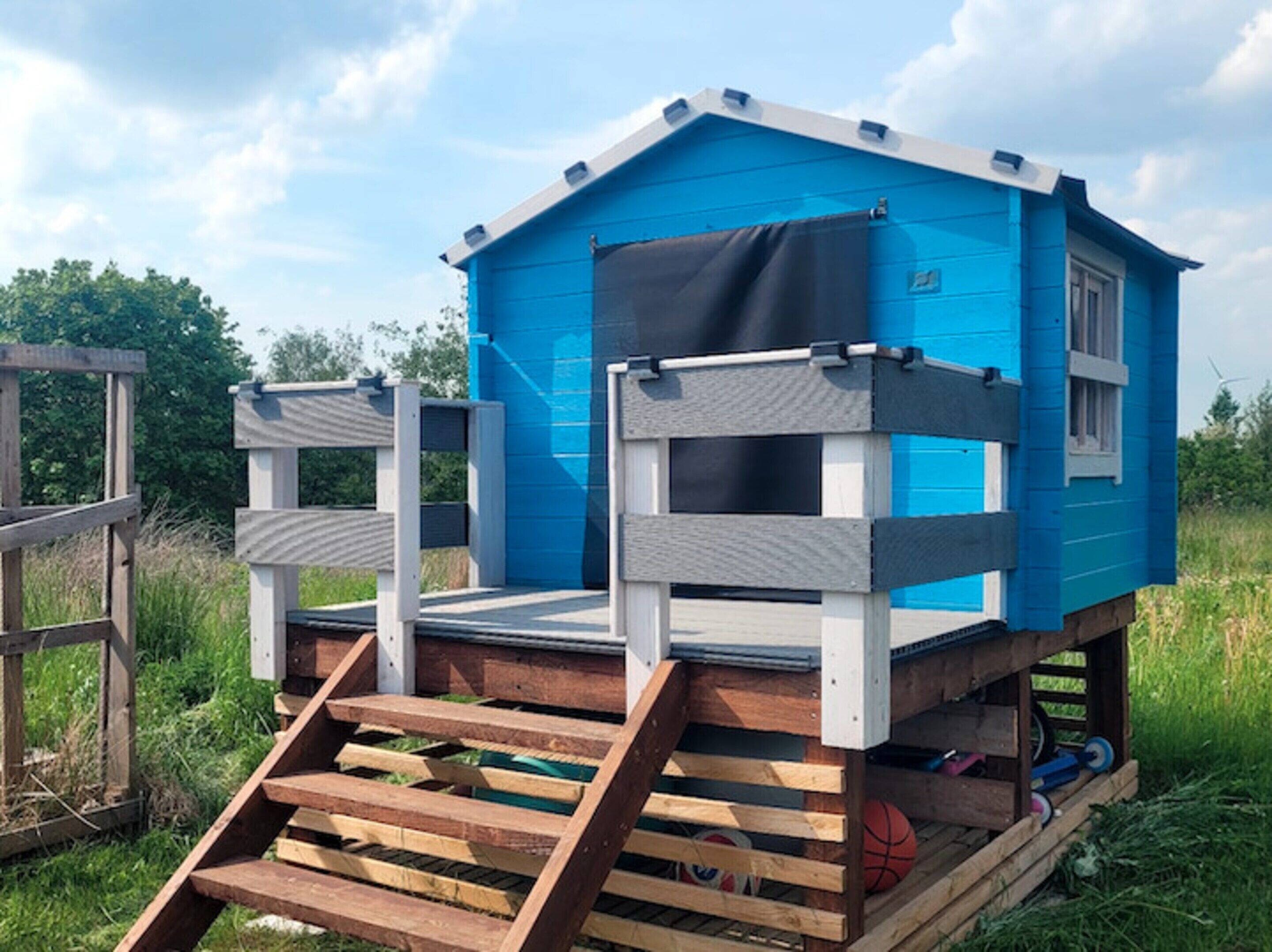 Ein blaues Holz-Spielhaus mit einer Holztreppe, umgeben von Gras und Bäumen.