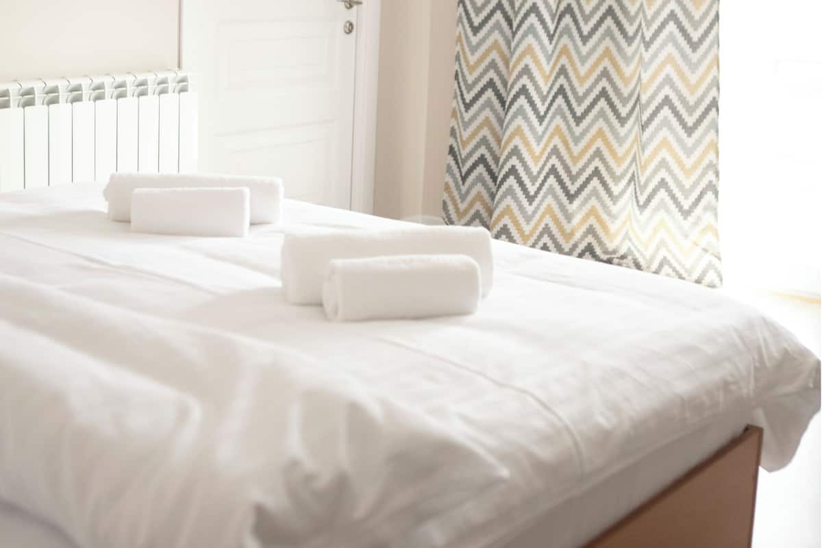 Un lit bien fait, avec du linge de lit blanc et deux serviettes enroulées, une porte blanche et un rideau à motifs chevrons.