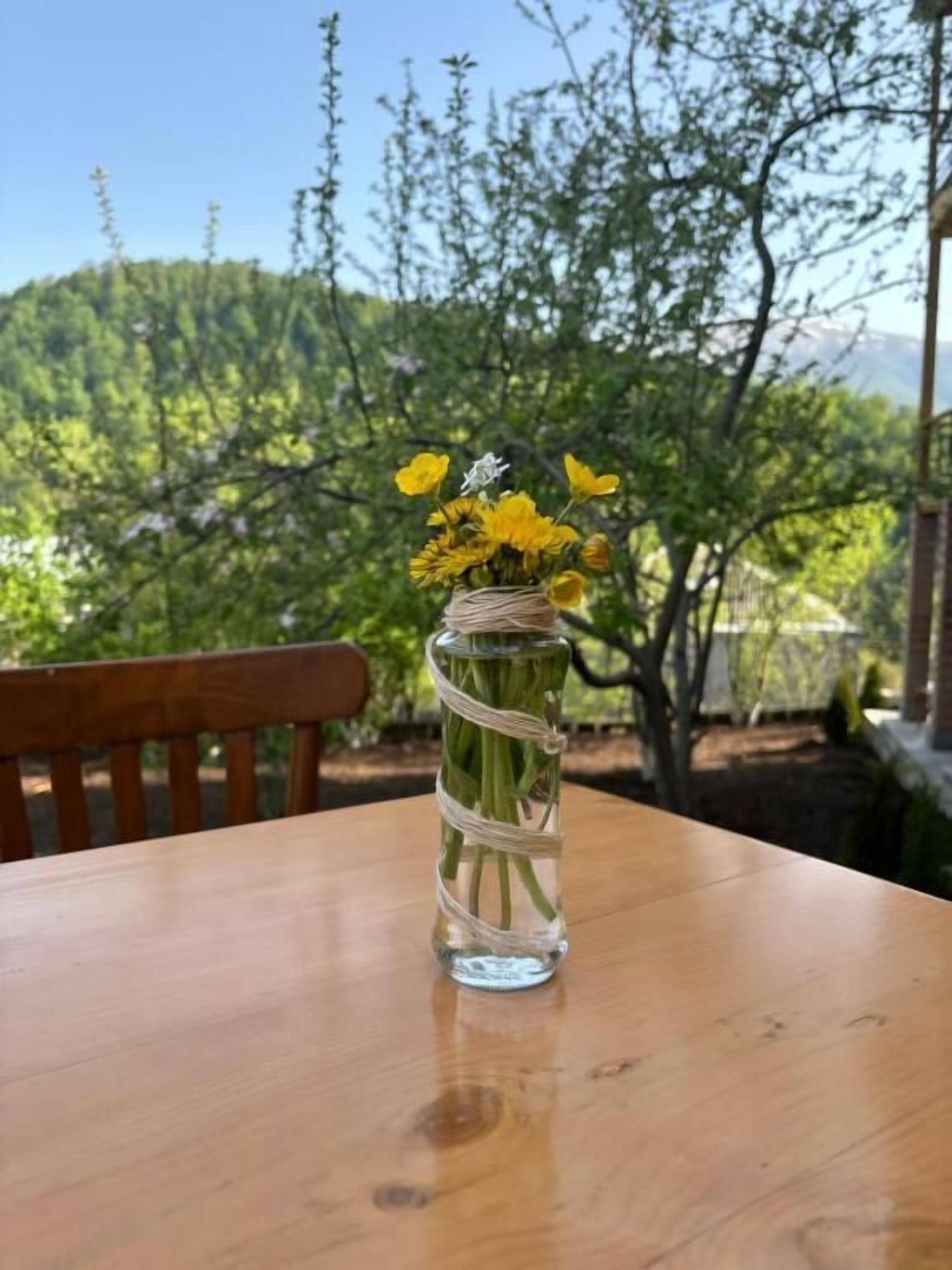 A clear glass vase with yellow flowers on a wooden table outdoors.