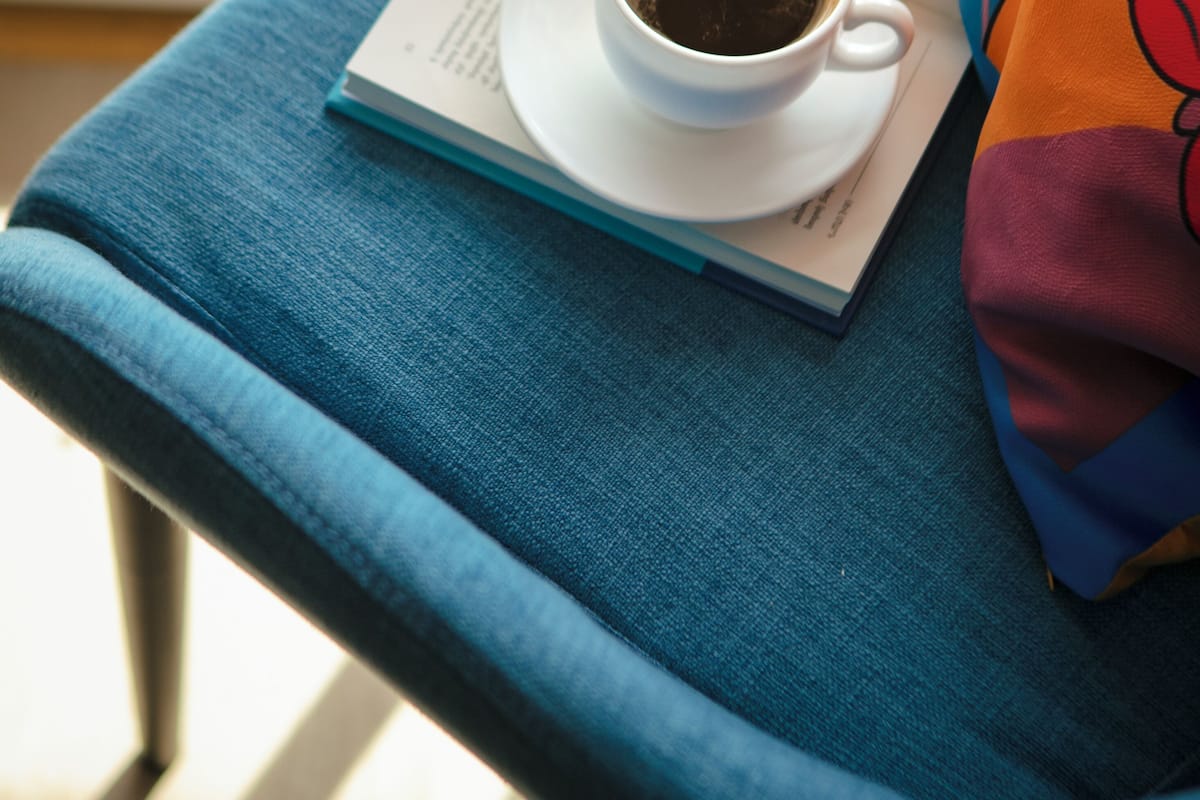Une tasse de café posée sur un livre, une chaise bleue et une couverture colorée.