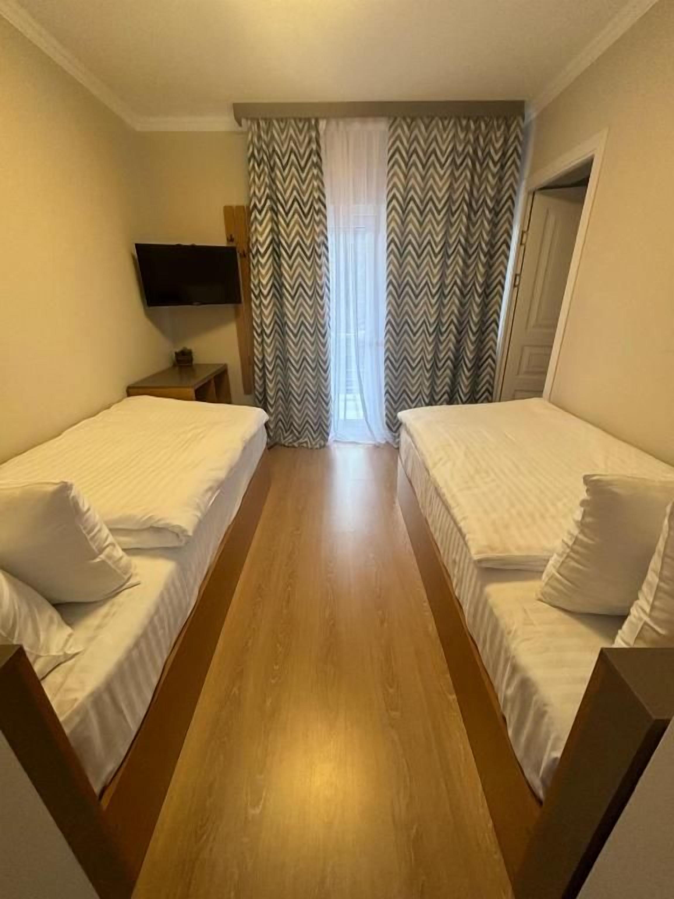 A hotel room with two single beds, a wooden floor, a television on the wall, and a window with curtains.
