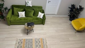 A green sofa with decorative pillows, a yellow armchair, a woven side table, and a patterned rug in a room with light wooden flooring and a white door. - RestArt Boutique Hotel (Dilijan)