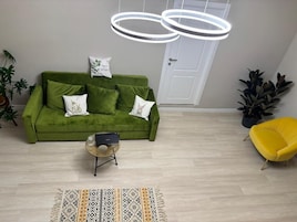 Un sofá verde con almohadones decorativos, un sillón amarillo, una mesita de noche tejida y una alfombra estampada en una habitación con suelo de madera clara y una puerta blanca.