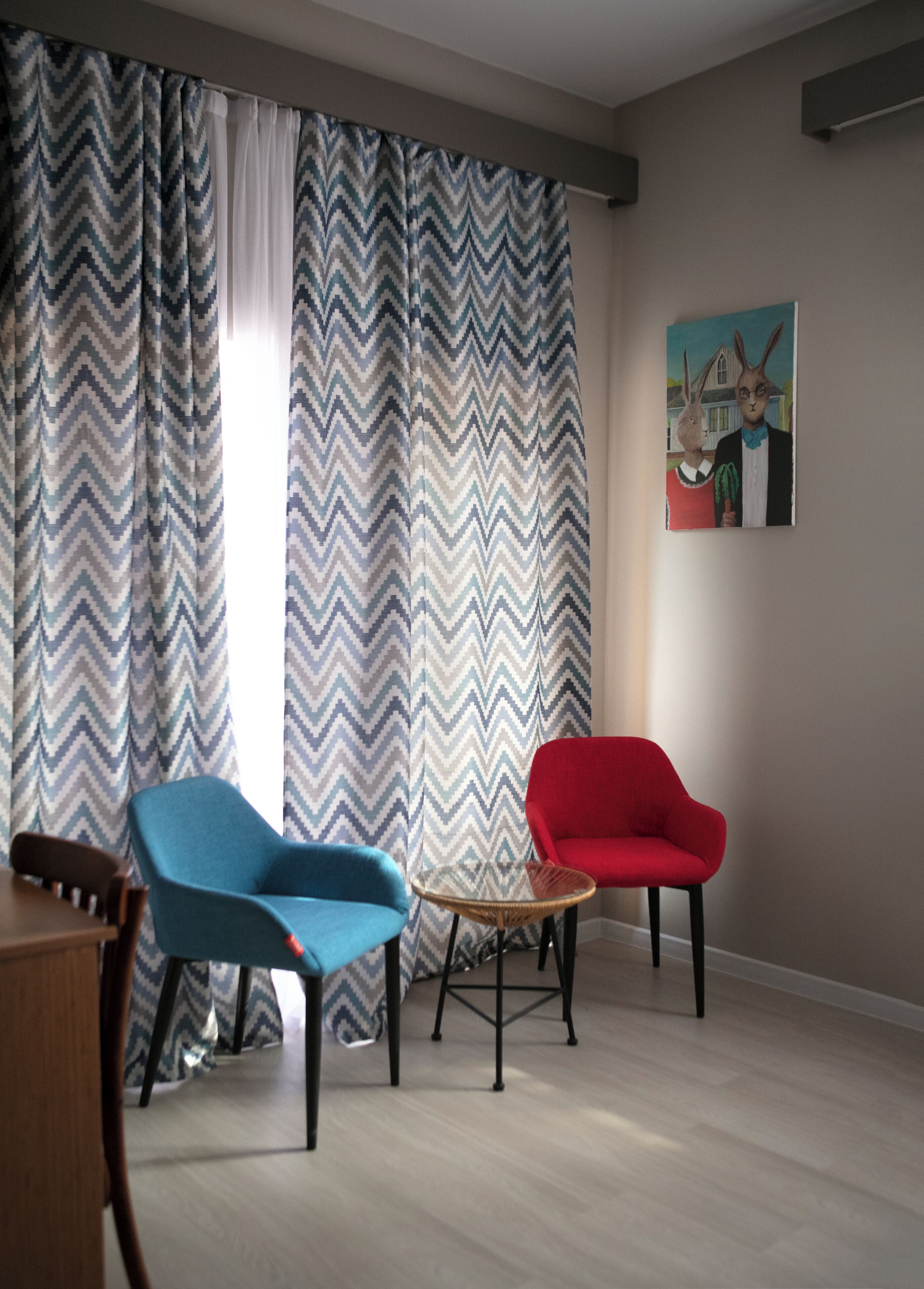 Une pièce avec un rideau à motifs en zigzag, un fauteuil bleu, un fauteuil rouge, une table en osier et un tableau accroché au mur.