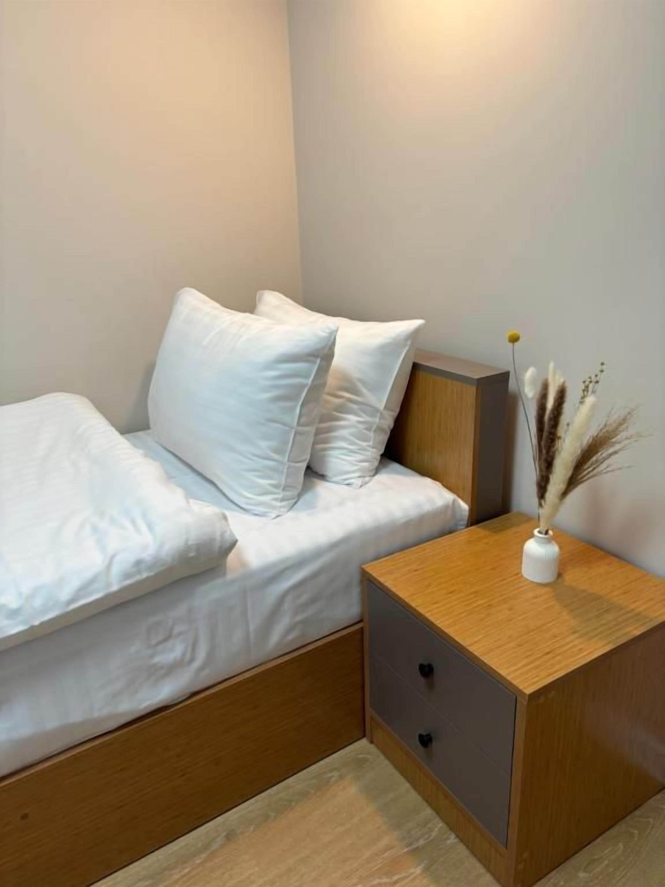 Una cama bien hecha con ropa blanca y dos almohadas, una mesita de noche de madera con un florero de plumas de pampa secas, y una lámpara de pared.