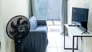Een ruime, goed verlichte kamer met een bed, een bureau met een computer, een ventilator en een raam met gordijnen.
