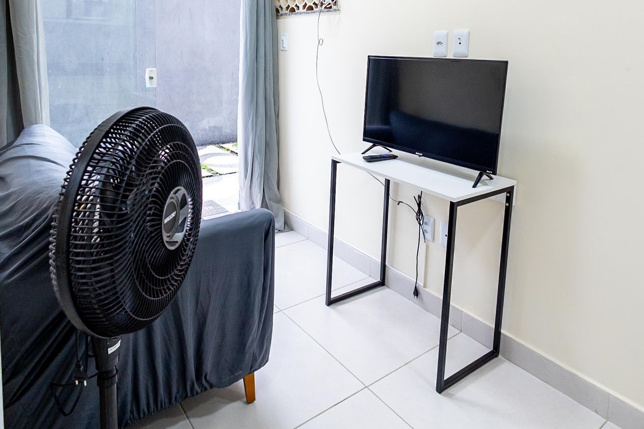 Una habitación con una cama, un ventilador de pie, un televisor sobre un mueble bajo y una ventana con cortinas.