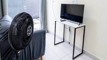 Una habitación con una cama, un ventilador de pie, un televisor sobre un mueble bajo y una ventana con cortinas.
