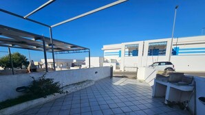 Una terrassa al terrat amb una pèrgola, un paviment de rajoles i un edifici blanc amb detalls blaus al fons.