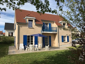 A two-story house with a red roof, blue shutters, and a balcony. There is a patio with white plastic chairs and a table.